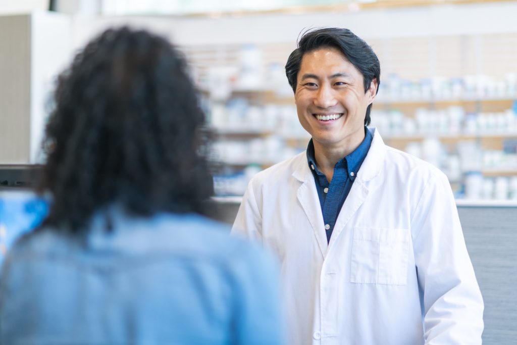 Pharmacien en blouse souriant à une patiente de dos au comptoir
